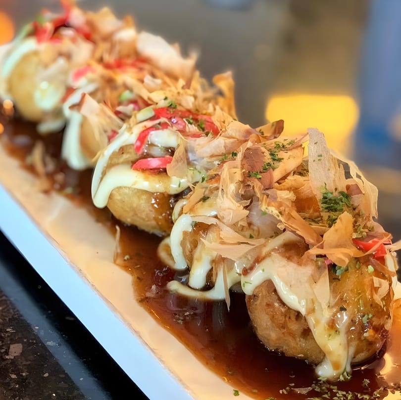 Takoyaki with mayonnaise, sauce, bonito flakes, and red ginger on a white rectangular plate.